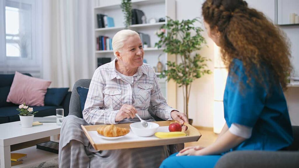 Older adult choosing clothing beside a caregiver in a bright assisted living room.