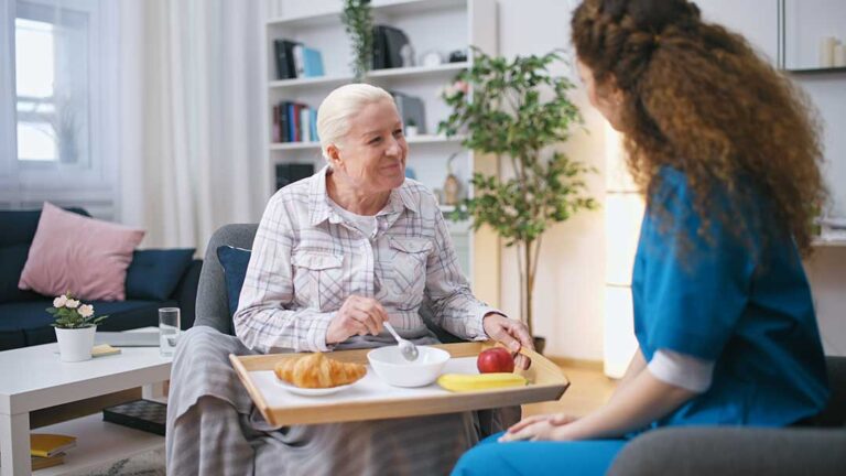 Older adult choosing clothing beside a caregiver in a bright assisted living room.